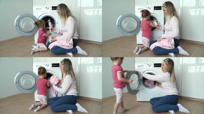 小微笑的3岁女儿的肖像和年轻的母亲把柔软的玩具放在洗衣机里洗。女孩关闭舱门，打开家用电器。