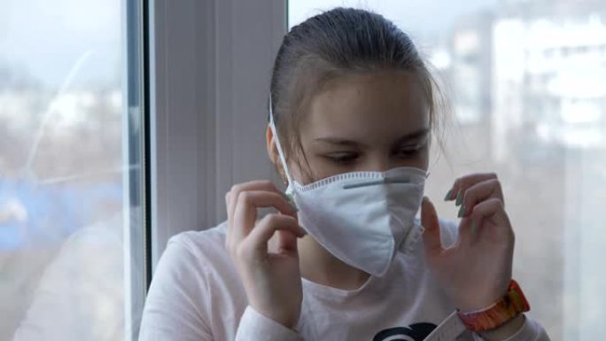 一个十几岁的女孩戴着防护医用口罩站在窗前。检疫生活。