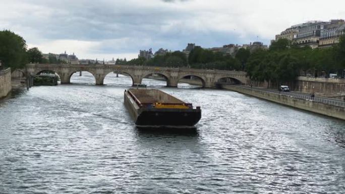 一艘货运驳船正沿着巴黎的塞纳河航行。她的波浪向两边发散。晴朗的天气，从上面拍摄。背景是一座桥。两侧是
