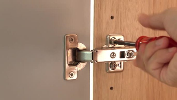 用螺丝刀在橱柜门上的抛光不锈钢门铰链上关闭男性手拧紧螺丝。