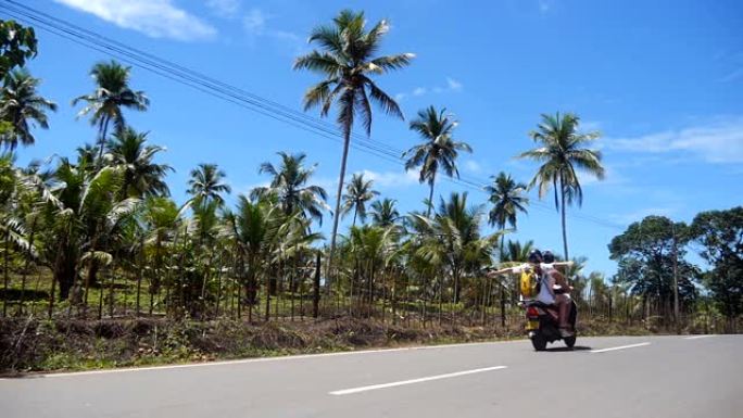 年轻女子背着背包，双臂伸出，与男友一起在棕榈树林立的道路上骑轻便摩托车。夫妇在热带岛屿上沿着沥青车道