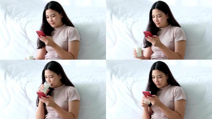 美丽的年轻亚洲女性坐在床边，在卧室的阳光下使用手机在线玩游戏