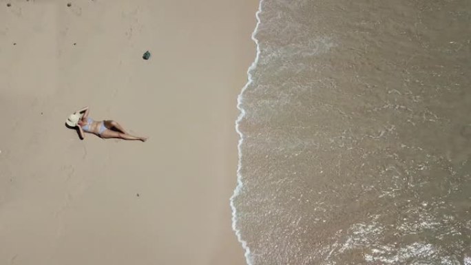 在印度尼西亚龙目岛粉红海滩的海边，一名戴着草帽的女孩躺在沙滩上的无人机拍摄。波浪轻轻地冲刷着海岸。女