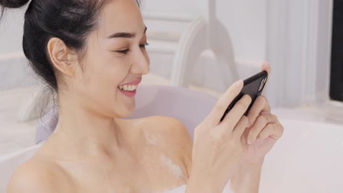 美丽的亚洲女人在浴室泡泡浴。她洗澡时带着快乐的情绪使用智能手机。