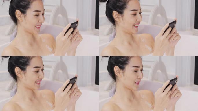 美丽的亚洲女人在浴室泡泡浴。她洗澡时带着快乐的情绪使用智能手机。