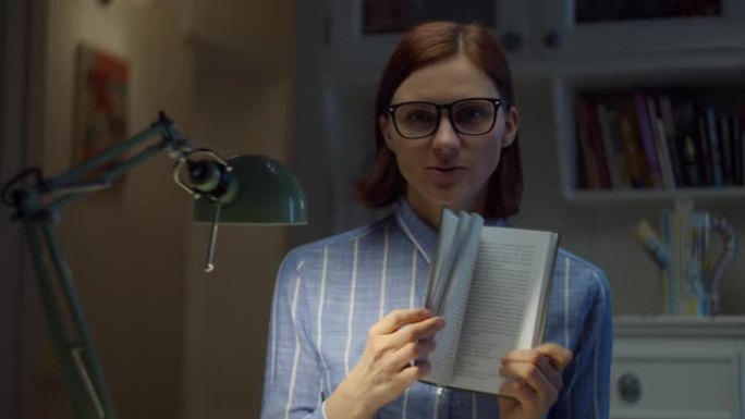 30多岁的女人戴着眼镜，展示纸质书，在家用相机解释。在线教育过程。老师说话的网络摄像头视图。
