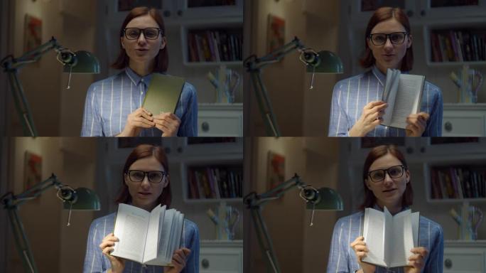 30多岁的女人戴着眼镜，展示纸质书，在家用相机解释。在线教育过程。老师说话的网络摄像头视图。