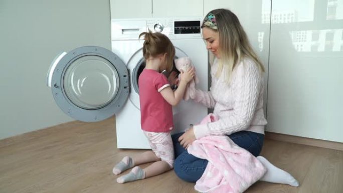 快乐的母亲带着小小的微笑，3岁的女儿把毛绒玩具放在洗衣机里洗。女孩关闭舱门，打开家用电器。家庭概念
