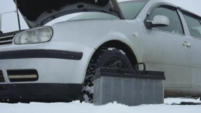 冬季降雪时汽车故障或问题。汽车电池躺在汽车旁边。引擎盖或引擎盖升起。典型的铅酸电池处于前台。慢动作