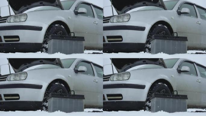 冬季降雪时汽车故障或问题。汽车电池躺在汽车旁边。引擎盖或引擎盖升起。典型的铅酸电池处于前台。慢动作