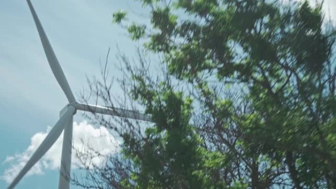 风力涡轮机的移动镜头，视图来自带有汽车的移动车辆。明亮的蓝天，有干净的白云，拍摄于夏日。绿色能源