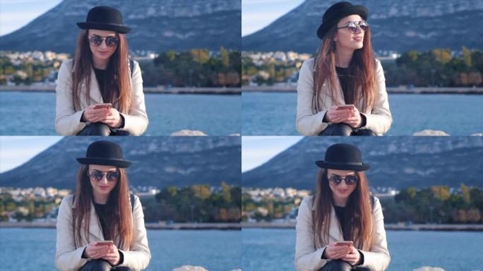 戴着黑帽子和眼镜的漂亮年轻女子在海边发短信