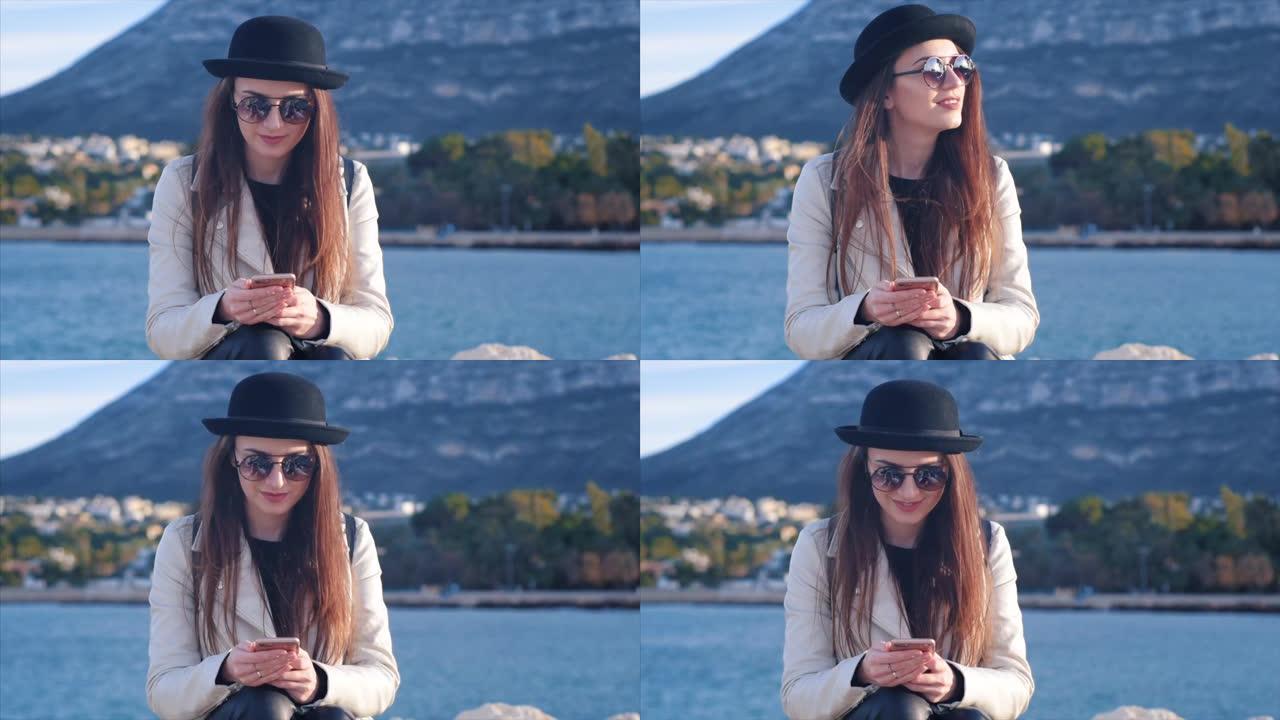 戴着黑帽子和眼镜的漂亮年轻女子在海边发短信