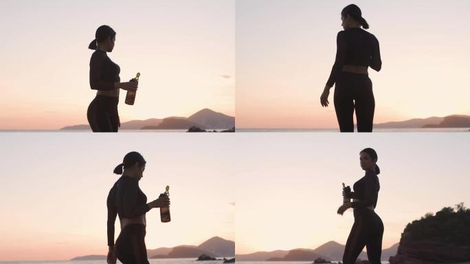 日落时在海滩上喝水的运动性感女人