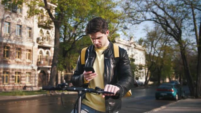 送货员的Poartrait与黄色袋装站在城市的电动踏板车，并使用智能手机找到客户的地址，慢动作