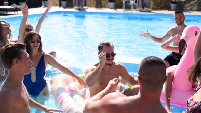 朋友在私人别墅游泳池聚会。在阳光明媚的日子里，穿着泳装的快乐年轻人在豪华度假胜地跳舞，用漂浮物和充气