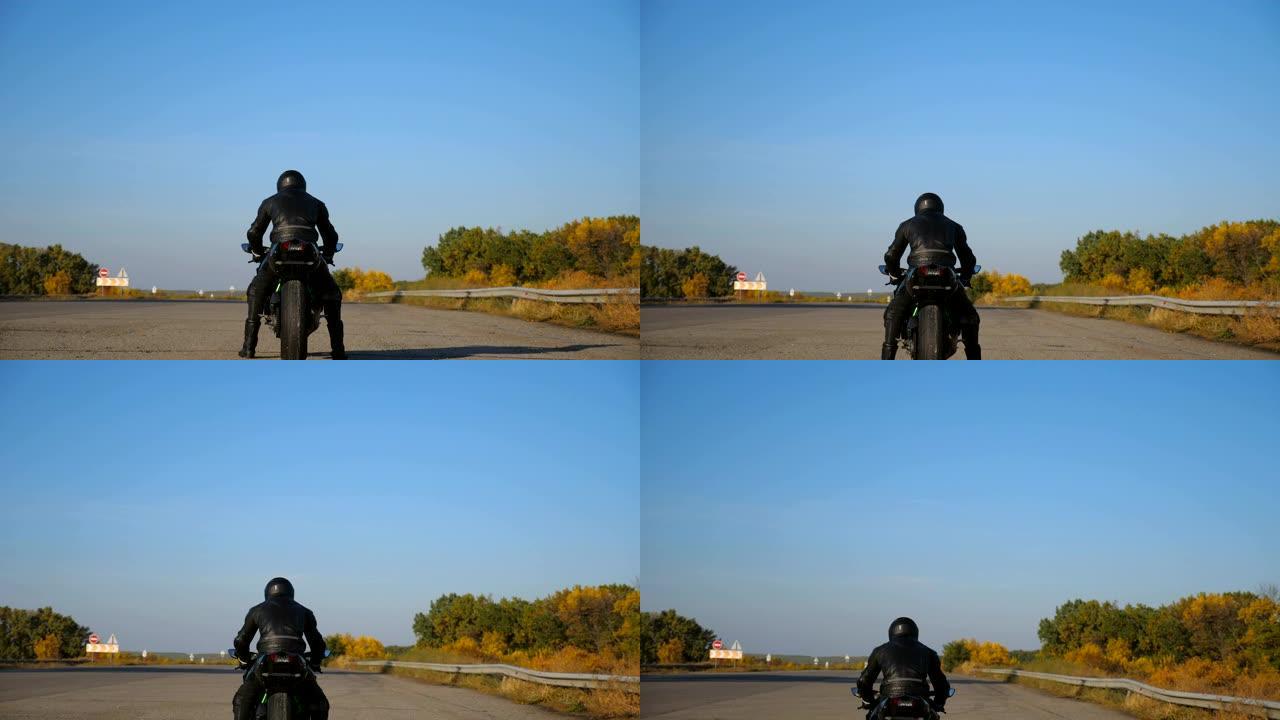 身穿头盔和皮夹克的无法识别的摩托车手的后视图正坐在高速公路上的摩托车上，准备骑行。男人要骑摩托车。自