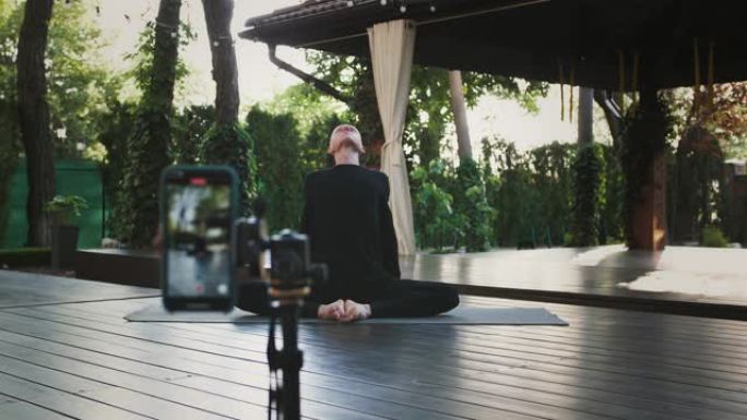 穿着黑色运动服的秃头男性坐在景观露台的垫子上表演瑜伽。使用三脚架上的智能手机拍摄自己的视频。特写