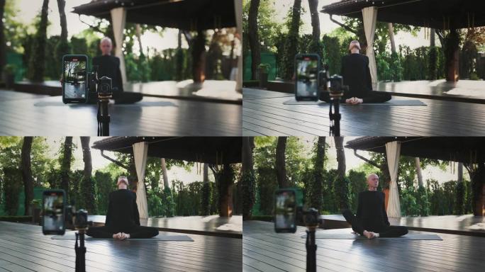 穿着黑色运动服的秃头男性坐在景观露台的垫子上表演瑜伽。使用三脚架上的智能手机拍摄自己的视频。特写