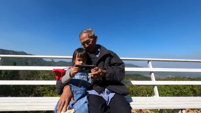 亚洲小女孩拿着智能手机与祖父坐在橙色种植园的白色木椅上自拍。