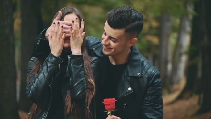 穿着黑色皮大衣的黑发女孩正在等她的男朋友。全黑的男孩闭上了女友的眼睛，献上一朵红玫瑰。背景模糊。慢动