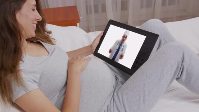 孕妇快乐的女人使用平板电脑和触摸她的腹部与她的医生在线交流。诊断咨询在线概念。妈妈在生孩子。怀孕。