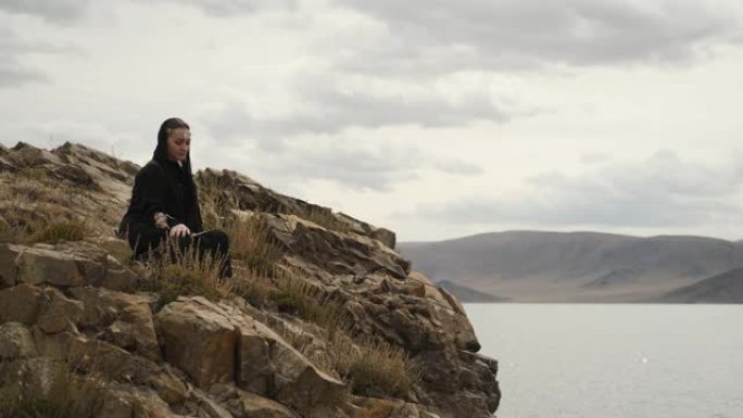 穿着黑色连衣裙的女人坐在山顶的悬崖边。大湖
