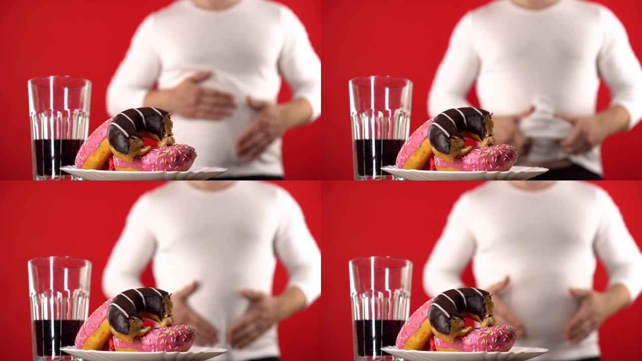 肥胖的概念。前景中的甜甜圈和苏打水，而在背景中，男人抚摸着他肥胖的腹部。甜蜜的情人暴饮暴食甜食。糟糕