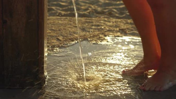 女人在沙滩上的沙滩淋浴下从腿上去除沙子，早晨的阳光照在背景中，细节上的水滴落在脚上