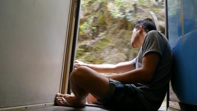 年轻人坐在火车开着的门附近，抽烟。男性游客乘坐铁路客运，俯瞰美丽的大自然。暑期旅游的概念。低视野
