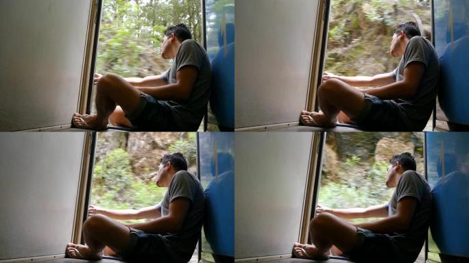 年轻人坐在火车开着的门附近，抽烟。男性游客乘坐铁路客运，俯瞰美丽的大自然。暑期旅游的概念。低视野