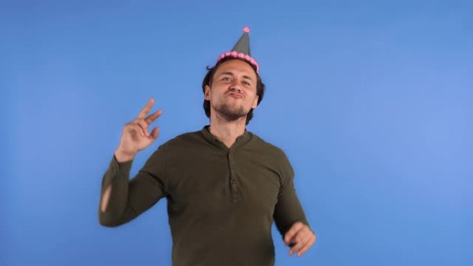 穿着党帽和卡其色衬衫的男人。他在蓝色背景上摆姿势时微笑，眨眼和跳舞。生日快乐庆祝。特写
