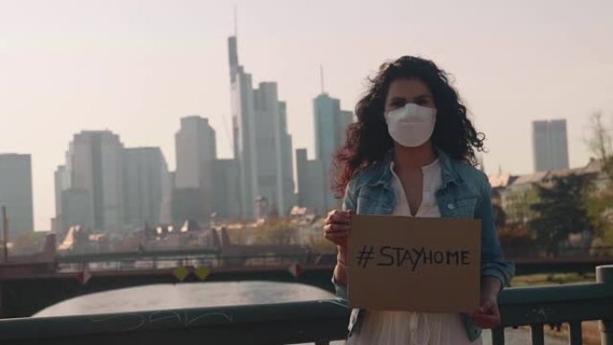 德国法兰克福，2020年4月8日年轻女性在天际线前手持 # stayhome标志，对着电晕病毒佩戴防