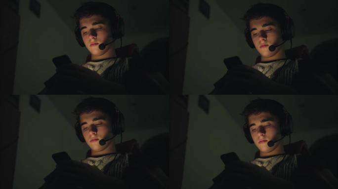 年轻的帅哥戴着带麦克风的耳机，使用智能手机，深夜坐在家里的电脑附近。带智能手机的发光脸