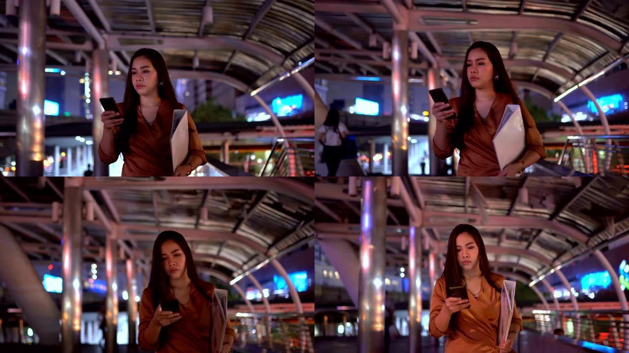 美丽的年轻亚洲女子使用智能彭走在夜城街道上