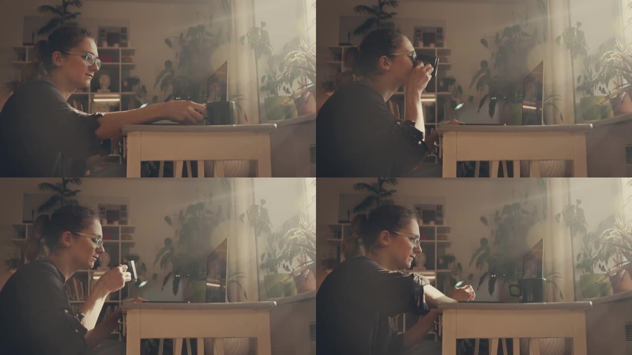 女孩自由职业者在家里的笔记本电脑上工作。她喝茶。窗户上刺眼的阳光。在书柜的背景下