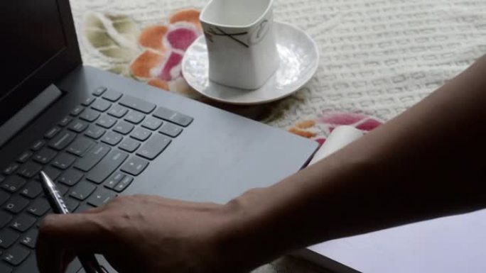 人手用笔记本电脑键盘打字。早餐咖啡用晨光笔和白纹纸笔记本，陶瓷杯盘放在办公室办公桌的顶部背景。