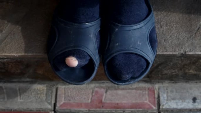 一个穿着破袜子和橡胶拖鞋的男人滑稽地移动了手指。男人穿着带洞的蓝色袜子。一个穷人的袜子。