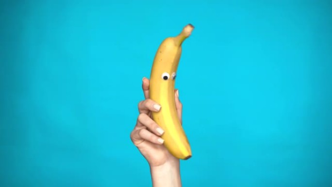 女人手里拿着眼睛的香蕉。香蕉跳入框架，在蓝色背景上环顾四周。女人手特写。