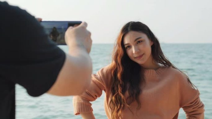 美丽的女人在海上对着镜头摆姿势。朋友使用智能手机拍摄带有海洋背景的照片。