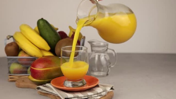 将醒酒器的果汁倒入玻璃杯中。一杯果汁。黄色清爽的夏季饮料。新鲜芒果汁、橙汁。桌子上的水果篮。厨房里有