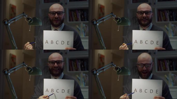 戴着眼镜的大胡子的30多岁男子在家里的相机上展示带有字母的纸张。在线教育过程。老师说话的网络摄像头视