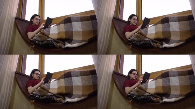 一名年轻女子正在平板电脑上发短信。一个女孩躺在窗边的窗台上，手里拿着平板电脑。窗外是明亮的一天。
