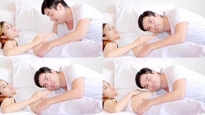 孕妇与丈夫躺在床上，试图听肚子里的声音说话，心情愉快。高加索男人和亚洲女人。年轻父母的概念。