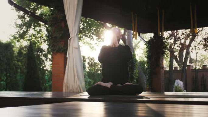 穿着黑色面具和运动服的强壮的家伙在花园露台的垫子和木地板上坐着莲花姿势表演瑜伽。冠状病毒大流行