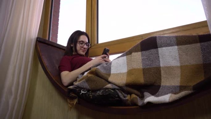 一名年轻女子正在打电话发短信。一个女孩躺在窗边的窗台上，手里拿着智能手机。窗外是明亮的一天。