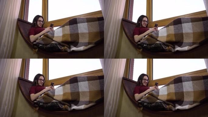 一名年轻女子正在打电话发短信。一个女孩躺在窗边的窗台上，手里拿着智能手机。窗外是明亮的一天。