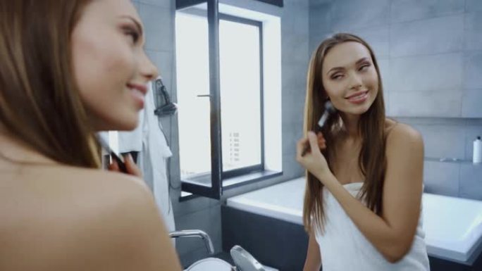 浴巾中的美丽年轻女子在镜子前使用化妆刷