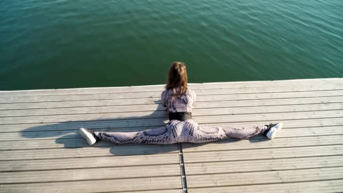 年轻苗条的女孩坐在一个美丽的湖旁的码头上。瑜伽和户外伸展运动。慢动作。顶视图。