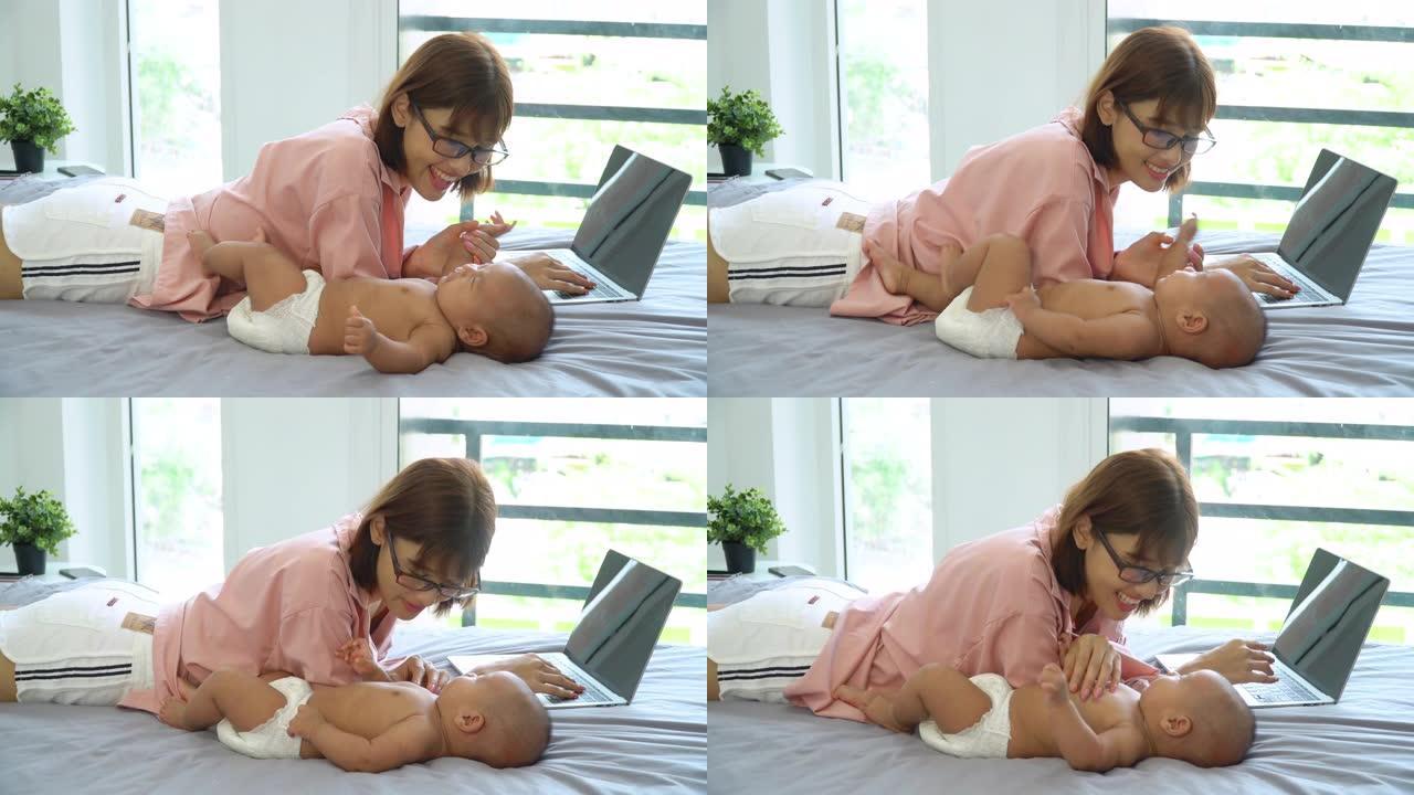 4k微笑的年轻美丽的亚洲女人母亲躺在床上，用笔记本电脑在家工作，和她可爱的新生女孩女儿玩耍。母婴关系